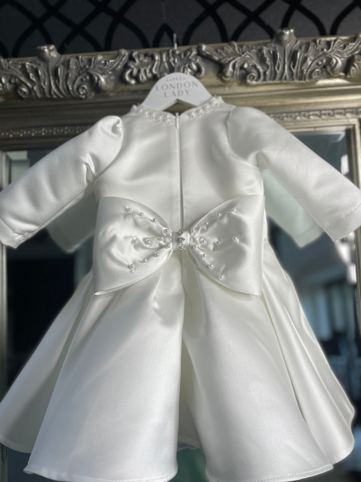 The stunning Satin Sofia Dress by Little London Lady. Handmade by Artisans. Pearl and beaded round neck with Ivory Satin Bodice. Long Satin Sleves or Puff Sleeve option. Large satin bow with pearl and diamond details. Perfect as a Baptism, Christening or naming ceromony dress. Size 6 months to 10 years old. custom sizing also available.